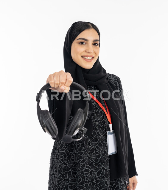 Using headphone technology, working in the field of music distribution, a Saudi woman who works in presenting radio programs, the concept of customer service, a portrait of a smiling Arab Gulf Emirati woman wearing a black abaya, wearing a personal identification card for employees and displaying her headset, white background.