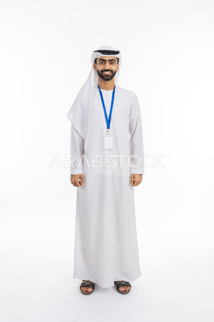 ارتداء بادج شخصي, النظر إلى الكاميرا بتعابير البهجة والفرح, الاهتمام بالمظهر الخارجي والأناقة الاماراتية, موك اب بطاقة تعريفية مفرغة باللون الابيض، بورتريه لرجل عربي خليجي إماراتي يرتدي الكندورة والغترة يقف بابتسامة, صورة الجسم بالكامل, خلفية بيضاء