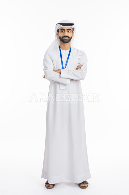 The concept of elegance and masculinity, attention to external appearance, standing with crossed hand gestures, portrait of an Emirati Gulf Arab man wearing a kandora and ghutra, looking at the camera and wearing an identification card with a blue ribbon, full-length body photo, white background