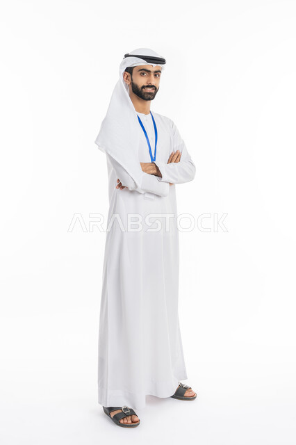 الوقوف بإيماءات أيدي متقاطعة, مفهوم الأناقة والرجولة, الاهتمام بالمظهر الخارجي, بورتريه لرجل عربي خليجي إماراتي يرتدي الكندورة والغترة ينظر إلى الكاميرا ويضع بطاقة شخصية تعريفية بشريط أزرق, صورة الجسم بالطول الكامل, خلفية بيضاء