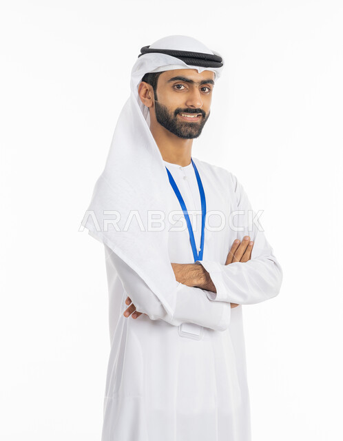 الاهتمام بالمظهر الخارجي, مفهوم الأناقة والرجولة, الوقوف بإيماءات أيدي متقاطعة, بورتريه لرجل عربي خليجي إماراتي مبتسم  يرتدي الكندورة والغترة ينظر إلى الكاميرا ويضع بطاقة شخصية تعريفية بشريط أزرق, خلفية بيضاء