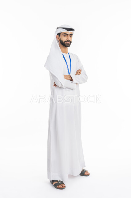 مفهوم الأناقة والرجولة, الاهتمام بالمظهر الخارجي, الوقوف بإيماءات أيدي متقاطعة, بورتريه لرجل عربي خليجي إماراتي يرتدي الكندورة والغترة ينظر إلى الكاميرا ويضع بطاقة شخصية تعريفية بشريط أزرق, صورة الجسم بالطول الكامل, خلفية بيضاء