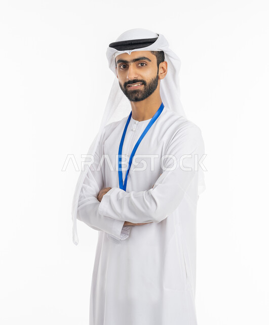 التقاط صور شخصية, الاهتمام بالمظهر الخارجي, مفهوم الأناقة والرجولة, الوقوف بإيماءات أيدي متقاطعة, بورتريه لرجل عربي خليجي إماراتي مبتسم يرتدي الكندورة والغترة ينظر إلى الكاميرا ويضع بطاقة شخصية تعريفية بشريط أزرق, خلفية بيضاء