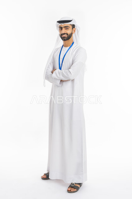 النظر إلى الكاميرا والوقوف بإيماءات أيدي متقاطعة, بورتريه لشاب عربي خليجي إماراتي مبتسم يرتدي الكندورة والغترة يضع هوية شخصية بشريط أزرق, ارتداء بادج شخصي, صورة الجسم بالطول الكامل, خلفية بيضاء