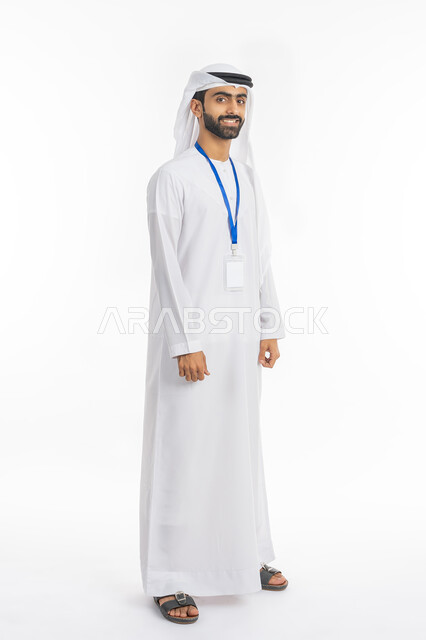 مفهوم الأناقة والرجولة والاهتمام بالمظهر الخارجي, كرت أبيض مستطيل فارغ, ارتداء بادج شخصي, بورتريه لرجل عربي خليجي إماراتي مبتسم يرتدي الكندورة والغترة ينظر إلى الكاميرا ويضع بطاقة شخصية تعريفية بشريط أزرق, صورة الجسم بالطول الكامل, خلفية بيضاء