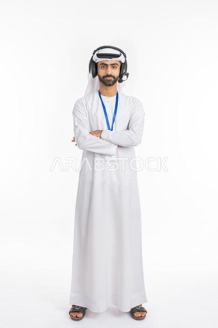 الإجابة على استفسارات الزبائن, مفهوم التقديم الاذاعي, الوقوف بإيماءات أيدي متقاطعة, بورتريه لموظف عربي خليجي إماراتي يرتدي الكندورة والغترة ينظر إلى الكاميرا, العمل في قسم الدعم الفني وخدمة العملاء, استخدام تقنية الهيدفون, صورة الجسم بالكامل, خلفية بيضاء