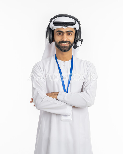 الوقوف بإيماءات أيدي متقاطعة, بورتريه لموظف عربي خليجي إماراتي مبتسم يرتدي الكندورة والغترة ينظر إلى الكاميرا, العمل في قسم الدعم الفني وخدمة العملاء, استخدام تقنية الهيدفون, الإجابة على استفسارات الزبائن, مفهوم التقديم الاذاعي, خلفية بيضاء