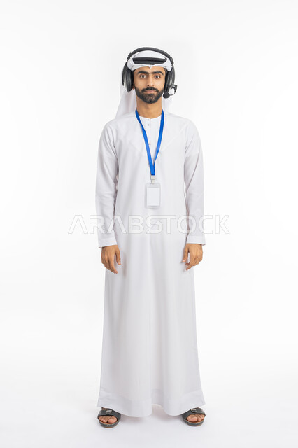 الوقوف باستقامة والنظر إلى الكاميرا, بورتريه لرجل عربي خليجي إماراتي يرتدي الكندورة والغترة الامارتية يستخدم تقنية الهيدفون, العمل في مجال خدمة العملاء, تقديم البرامج الاذاعية, موك أب أبيض مستطيل فارغ, صورة الجسم بالطول الكامل, خلفية بيضاء