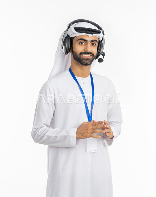 برامج التحدث عن بعد, استخدام تقنية الهيدفون, الرد على اسئلة العملاء بإيماءات البشاشة, بورتريه لرجل عربي خليجي إماراتي مبتسم يرتدي الكندورة والغترة الاماراتية يقف بثقة بالنفس, ارتداء بادج شخصي, العمل في قسم الدعم الفني, خلفية بيضاء
