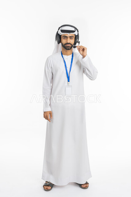 Standing straight and looking at the camera, concept of working in the field of customer service, portrait of an Emirati Gulf Arab man wearing a kandora and ghutra with his hands on the headset, use of headphone technology, full-length body portrait, white background