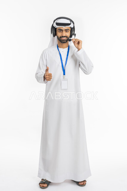 النظر إلى الكاميرا بإيماءات الثقة بالنفس, تقديم البرامج الاذاعية, استخدام تقنية الهيدفون, ارتداء بادج شخصي, مفهوم خدمة العملاء, بورتريه لموظف عربي خليجي إماراتي يرتدي الكندورة والغترة يرفع اصبعه بعلامة الجودة والاميتاز, صورة الجسم بالطول الكامل, خلفية بيضاء