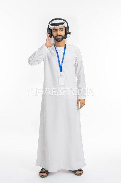 Presenting radio programs, using headphone technology, wearing a personal badge with a blue ribbon, the concept of customer service and responding to customer inquiries, a portrait of an Arab Gulf-Emirati employee wearing a kandora and ghutra looking at the camera with serious gestures, full-length body portrait, white background
