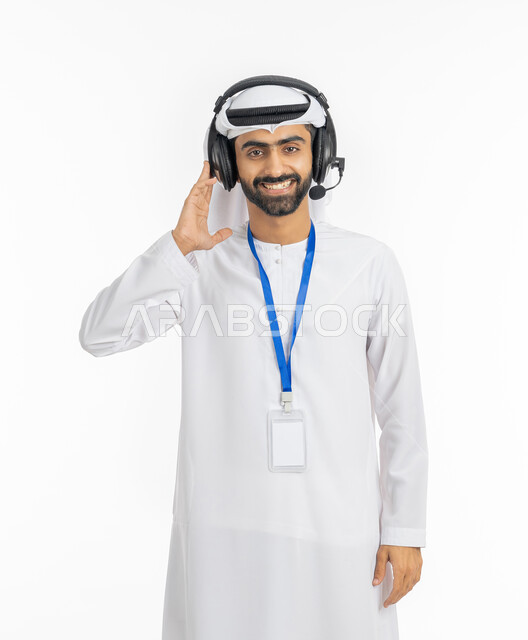 ارتداء بادج شخصي بشريط أزرق, مفهوم خدمة العملاء والرد على استفسارات الزبائن, بورتريه لموظف عربي خليجي إماراتي مبتسم يرتدي الكندورة والغترة ينظر إلى الكاميرا بإيماءات السرور, استخدام تقنية الهيدفون, تقديم البرامج الاذاعية, خلفية بيضاء