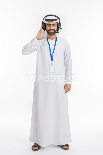 مفهوم خدمة العملاء والرد على استفسارات الزبائن, بورتريه لموظف عربي خليجي إماراتي مبتسم يرتدي الكندورة والغترة ينظر إلى الكاميرا بإيماءات السرور, استخدام تقنية الهيدفون, تقديم البرامج الاذاعية, ارتداء بادج شخصي بشريط أزرق, صورة الجسم بالطول الكامل, خلفية بيضاء