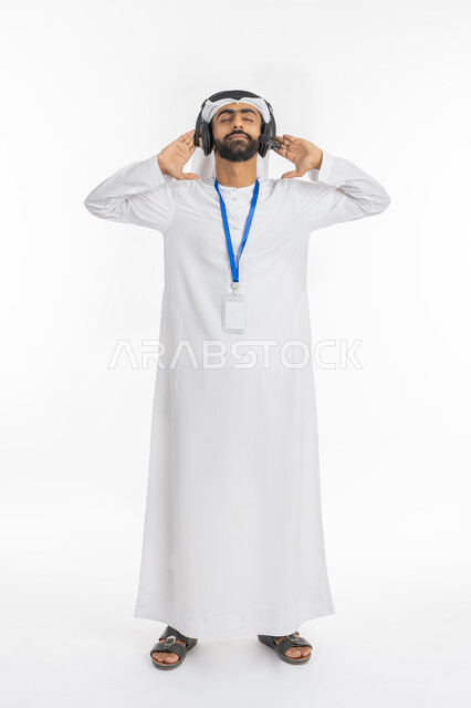 التفاعل والطرب والإصغاء الموسيقي, بورتريه لرجل عربي خليجي سعودي يرتدي الكندورة والغترة يضع يديه على الهيدفون بإيماءات الاندماج لسماع الموسيقى, ارتداء بادج شخصي, بطاقة تعريفية باللن الأبيض مستطيلة مفرغة, صورة الجسم بالطول الكامل, خلفية بيضاء