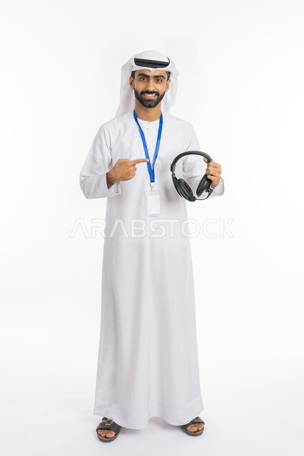 العمل في مجال خدمة العملاء والرد على اسئلة الزبائن, تقديم البرامج الاذاعية, بورتريه لشاب عربي خليجي إماراتي يرتدي الكندورة والغترة الاماراتية يشير بإصبعه إلى سماعة الرأس, استخدام تقنية الهيدفون, بطاقة شخصية تعريفية بشريط أزرق, صورة الجسم بالكامل, خلفية بيضاء