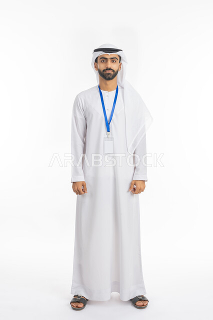 الوقوف باستقامة والنظر إلى الكاميرا بإيماءات الجدية, بورتريه لشاب عربي خليجي إماراتي يرتدي الكندورة والغترة يضع هوية شخصية بشريط أزرق, ارتداء بادج شخصي, كرت مستطيل الشكل أبيض اللون, صورة الجسم بالطول الكامل, خلفية بيضاء