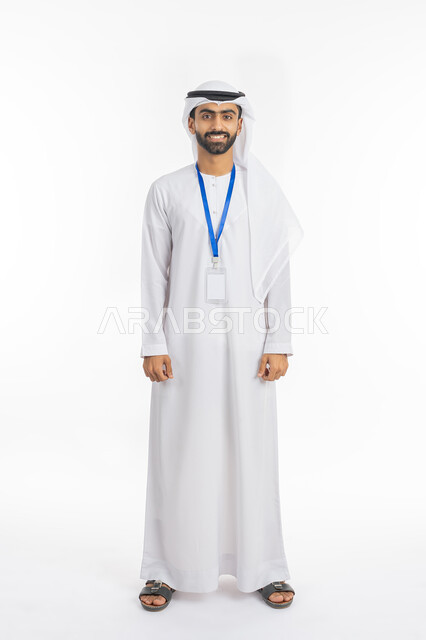 مفهوم الرجولة والاهتمام بالمظهر الخارجي, موك اب بطاقة تعريفية مفرغة باللون الابيض, النظر إلى الكاميرا بتعابير البهجة والفرح، بورتريه لرجل عربي خليجي إماراتي يرتدي الكندورة والغترة الاماراتية يقف بابتسامة, ارتداء بادج شخصي, صورة الجسم بالطول الكامل, خلفية بيضاء