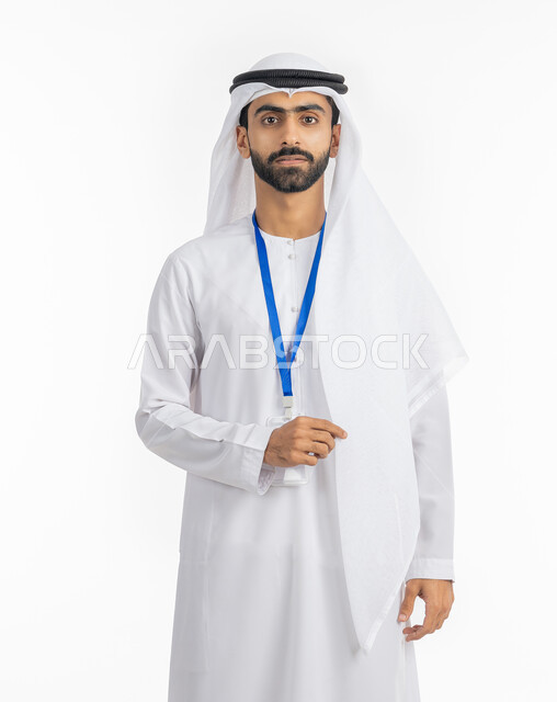 الوقوف بإيماءات الجدية والقوة, مفهوم الرجولة والاهتمام بالمظهر الخارجي, بورتريه لرجل عربي خليجي إماراتي يرتدي الكندورة والغترة ينظر إلى الكاميرا ويضع بطاقة شخصية تعريفية بشريط أزرق, خلفية بيضاء