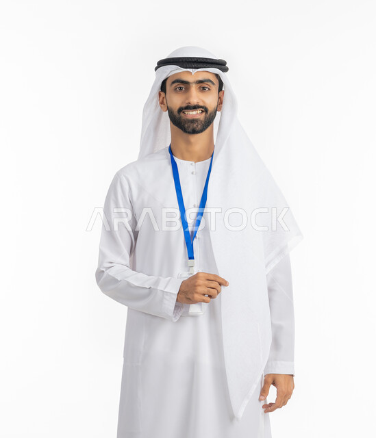 مفهوم الرجولة والاهتمام بالمظهر الخارجي, الوقوف بإيماءات البشاشة والسرور, بورتريه لرجل عربي خليجي إماراتي يرتدي الكندورة والغترة ينظر إلى الكاميرا ويضع بطاقة شخصية تعريفية بشريط أزرق, خلفية بيضاء