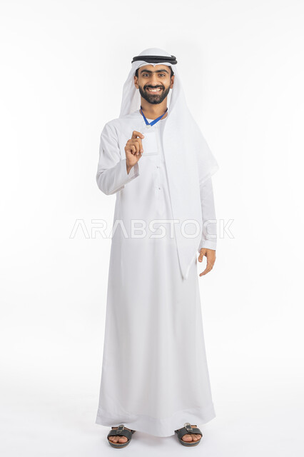 بطاقة تعريفية شخصية باسم الموظف, عرض كرت أبيض مستطيل فارغ, الوقوف والنظر إلى الكاميرا بإيماءات السرور, بورتريه لرجل أعمال عربي خليجي إماراتي مبتسم يرتدي الكندورة والغترة يضع هوية شخصية, ارتداء بادج شخصي, صورة الجسم بالطول الكامل, خلفية بيضاء
