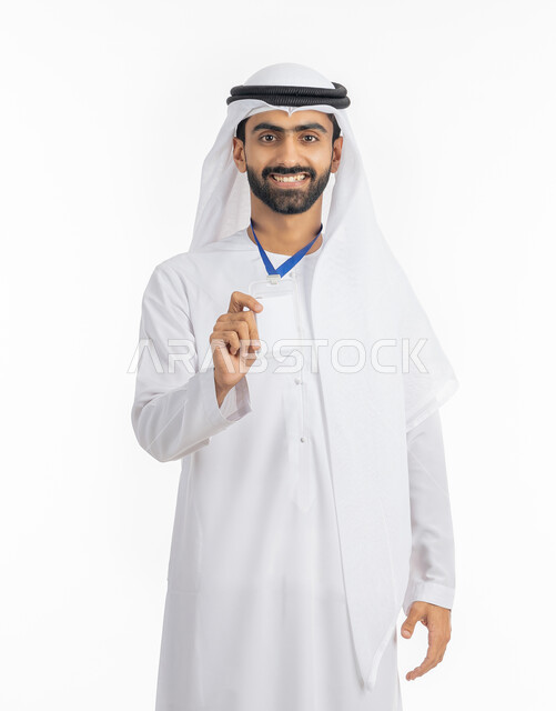 عرض كرت أبيض مستطيل فارغ, الوقوف والنظر إلى الكاميرا بإيماءات السرور, بورتريه لرجل أعمال عربي خليجي إماراتي مبتسم يرتدي الكندورة والغترة يضع هوية شخصية, ارتداء بادج شخصي, بطاقة تعريفية شخصية باسم الموظف, خلفية بيضاء