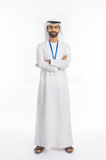 Caring about external appearance, the concept of elegance and masculinity, standing with crossed hand gestures, portrait of an Emirati Gulf Arab man wearing a kandora and ghutra, looking at the camera and wearing an identification card with a blue ribbon, full-length body photo, white background
