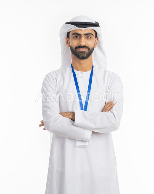 مفهوم الرجولة والاهتمام بالمظهر الخارجي, الوقوف بإيماءات أيدي متقاطعة, بورتريه لرجل عربي خليجي إماراتي يرتدي الكندورة والغترة ينظر إلى الكاميرا ويضع بطاقة شخصية تعريفية بشريط أزرق, خلفية بيضاء