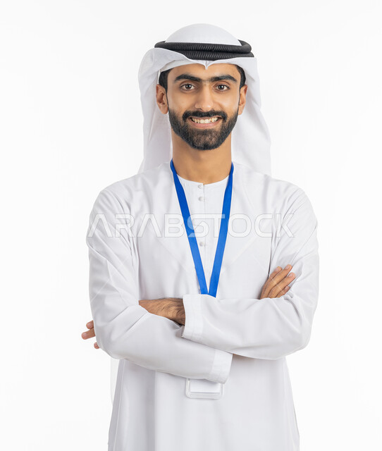 الوقوف بإيماءات أيدي متقاطعة, بورتريه لرجل عربي خليجي إماراتي يرتدي الكندورة والغترة ينظر إلى الكاميرا بابتسامة ويضع بطاقة شخصية تعريفية بشريط أزرق, مفهوم الرجولة والاهتمام بالمظهر الخارجي, خلفية بيضاء