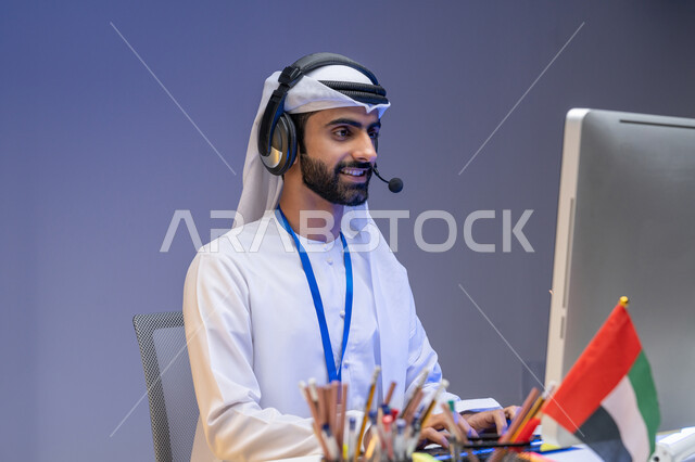 وظائف ومهن مكتبية, الاعتزاز بالانتماء لدولة الإمارات, العمل في مجال خدمة العملاء, الرد على استفسارات الزبائن, بورتريه مقرب لموظف عربي خليجي إماراتي يرتدي الكندورة والغترة يضع سماعة الاتصال يعمل على الحاسوب في المكتب بإيماءات الاندماج, خلفية ملونة