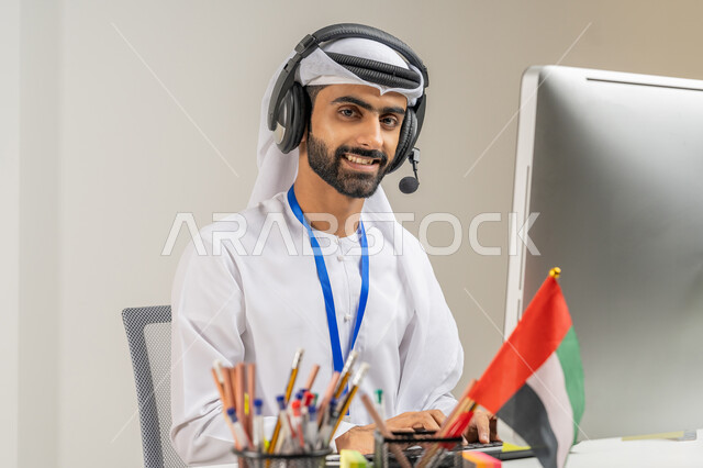 العمل في مجال خدمة العملاء, النظر إلى الكاميرا بتعابير البهجة والسرور, الفخر والاعتزاز بالانتماء لدولة الإمارات, بورتريه مقرب لموظف عربي خليجي إماراتي يرتدي الكندورة والغترة يضع سماعة الاتصال يعمل على الحاسوب في المكتب, الرد على استفسارات الزبائن, خلفية رمادية