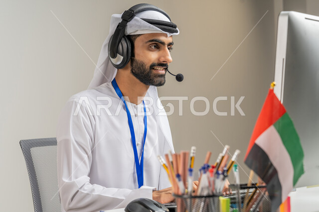 الاعتزاز بالانتماء لدولة الإمارات, العمل في مجال خدمة العملاء, الرد على استفسارات الزبائن, بورتريه مقرب لموظف عربي خليجي إماراتي يرتدي الكندورة والغترة يضع سماعة الاتصال يعمل على الحاسوب في المكتب بإيماءات الاندماج, خلفية رمادية