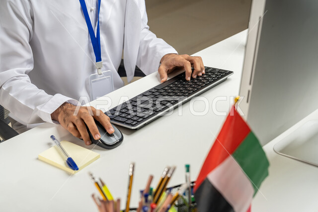 مهن ووظائف مكتبية, النقر والكتابة على لوحة المفاتيح, استخدام جهاز الحاسوب في العمل، إدارة شؤون الأعمال وتنظيمها عبر الاجهزة التقنية, صورة مقربة لموظف عربي خليجي إماراتي يرتدي الكندورة يجلس في المكتب, تطوير استراتيجية العمل في الشركات الاماراتية, خلفية رمادية