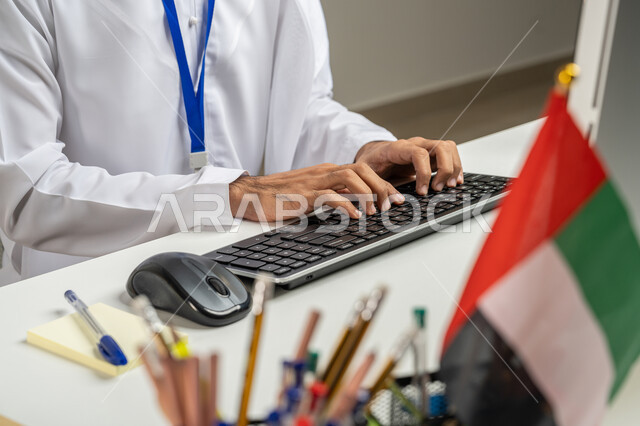 النقر والكتابة على لوحة المفاتيح, استخدام جهاز الحاسوب في العمل، إدارة شؤون الأعمال وتنظيمها عبر الاجهزة التقنية, صورة مقربة لموظف عربي خليجي إماراتي يرتدي الكندورة يجلس في المكتب, مهن ووظائف مكتبية, تطوير استراتيجية العمل في الشركات الاماراتية, خلفية رمادية
