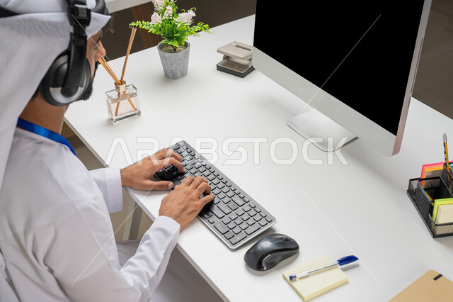 اعداد وتقديم البرامج الإذاعية، الإجابة على أسئلة العملاء، دمج التقنيات الحديثة في الحياة العملية, بورتريه مقرب من الجانب لموظف عربي خليجي اماراتي يرتدي الكندورة يضع سماعة التواصل يعمل في مجال خدمة الزبائن، أداء المهام عبر جهاز الحاسوب في المكتب, خلفية رمادية