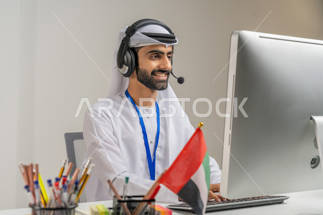 العمل في مجال خدمة العملاء, الرد على استفسارات الزبائن, الفخر والاعتزاز بالانتماء لدولة الإمارات, بورتريه مقرب لموظف عربي خليجي إماراتي يرتدي الكندورة والغترة يضع سماعة الاتصال يعمل على الحاسوب في المكتب بإيماءات الاندماج, خلفية رمادية