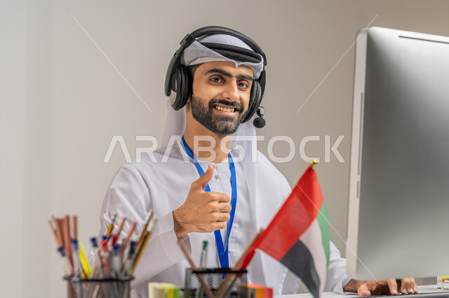 الفخر والاعتزاز بالانتماء لدولة الإمارات, بورتريه مقرب لموظف عربي خليجي إماراتي يرتدي الكندورة والغترة يرفع إصبعه بإيماءات الجودة والامتياز في المكتب, استخدام تقنية الهيدفون, العمل في مجال خدمة العملاء, دمج التقنيات المتطورة في الحياة العملية، خلفية رمادية