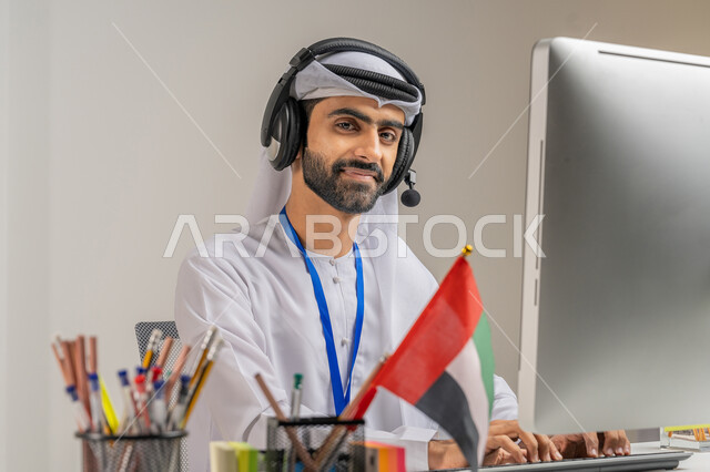 النظر إلى الكاميرا بتعابير البهجة والسرور, الفخر والاعتزاز بالانتماء لدولة الإمارات, بورتريه مقرب لموظف عربي خليجي إماراتي يرتدي الكندورة والغترة يضع سماعة الاتصال يعمل على الحاسوب في المكتب, العمل في مجال خدمة العملاء, الرد على استفسارات الزبائن, خلفية رمادية
