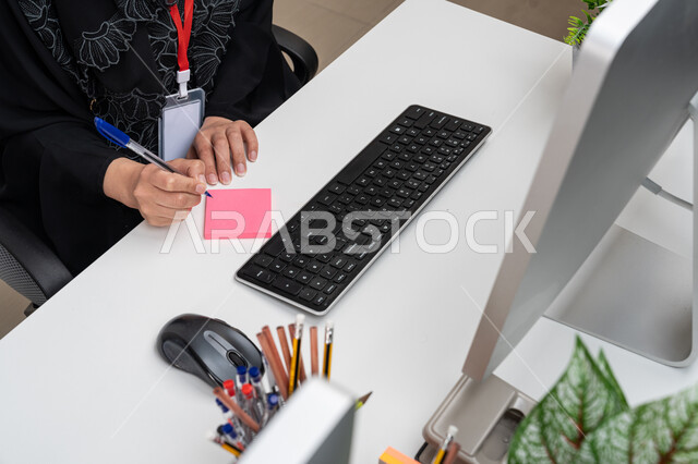 استخدام الأجهزة والتقنيات الحديثة والمتطورة, قسم خدمة العملاء, مشاركة النساء في الوظائف المكتبية، صورة مقربة من الأعلى لموظفة عربية خليجية إماراية ترتدي العباءة السوداء تجلس في المكتب وتقوم بكتابة الملاحظات على الورقة, أداء وإدارة المهام عبر جهاز الحاسوب