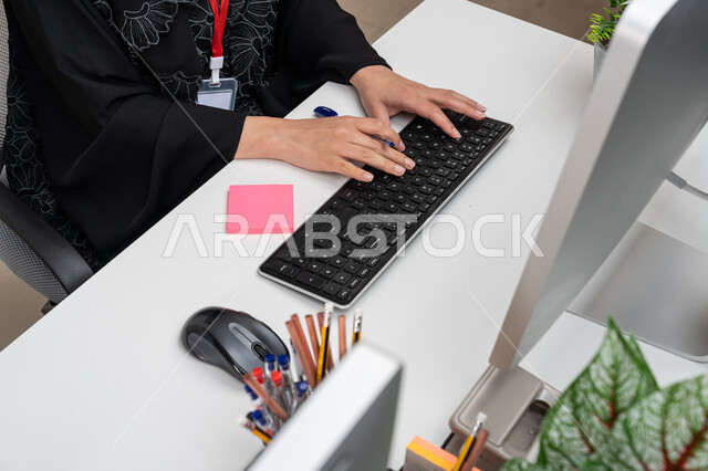 أداء وإنجاز المهام عبر جهاز الحاسوب, استخدام الأجهزة والتقنيات الحديثة والمتطورة, إمرأة سعودية تعمل في قسم خدمة العملاء, مشاركة النساء في الوظائف المكتبية، صورة من الأعلى لموظفة عربية خليجية إماراتية ترتدي العباءة السوداء تجلس في المكتب