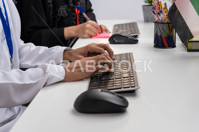 Using a computer at work, managing and organizing business affairs through technical devices, a close-up picture from the side of an Arab Gulf Emirati employee wearing a kandora sitting in the office, office professions and jobs, developing a work strategy in Emirati companies, clicking and typing on the keyboard