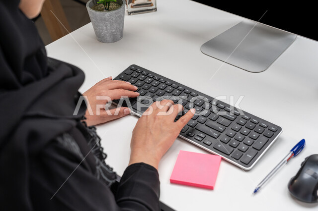 مفهوم خدمة العملاء، وظائف ومهن مكتبية، صورة مقربة من الخلف لموظفة عربية خليجية إماراتية محجبة ترتدي العباءة السوداء تجلس في مقر الشركة، إمرأة سعودية تعمل على جهاز الحاسوب، استخدام الأجهزة والتقنيات الحديثة والمتطورة