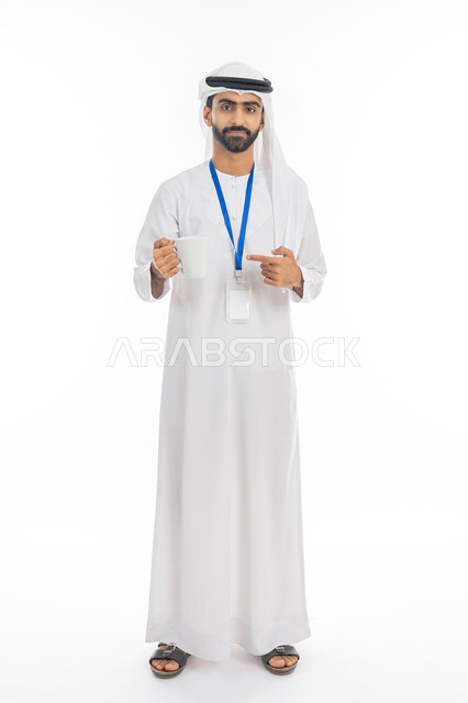 الوقوف باستقامة والنظر إلى الكاميرا, ارتداء بادج شخصي, بورتريه لرجل عربي خليجي إماراتي يرتدي الكندورة والغترة يضع كرت أبيض مستطيل فارغ بشريط أزرق ويقوم بالإشارة إلى كوب للمشروبات الساخنة, تأثير القهوة على صحة الدماغ، صورة الجسم بالطول الكامل, خلفية بيضاء