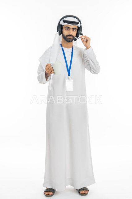 التواصل مع الزبائن لحل المشكلات في مكاتب خدمة العملاء، الاجابة على الاستفسارات والاستعلامات, بورتريه لرجل عربي خليجي إماراتي يرتدي الكندورة والغترة يضع الهيدفون ويمسك كوب من القهوة، دمج التقنيات المتطورة مع العمل, صورة الجسم بالطول الكامل, خلفية بيضاء