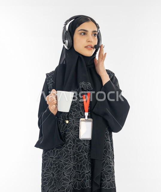 Integration and focus in performing work tasks, the concept of customer service, women’s jobs and professions, developing a business strategy, a portrait of an Arab Gulf Emirati woman wearing an abaya and a headset answering the customer’s questions, a Saudi woman holding a cup of coffee, white background