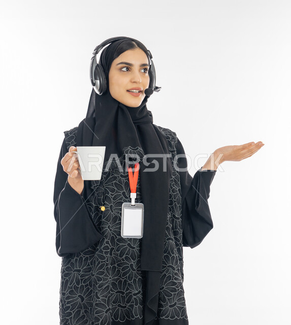 استفسار العميل عن المعلومات عبر الاتصال بمكاتب خدمة العملاء, بورتريه لإمرأة عربية خليجية اماراتية ترتدي العباءة السوداء تضع سماعة التواصل وتعمل في المجال المكتبي, سيدة سعودية تمسك كوب من القهوة وترفع يديها بإيماءات النقاش مع الزبون, خلفية بيضاء