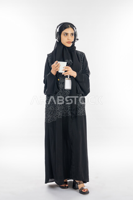 الاجابة على الاسئلة والتواصل مع العملاء, بورتريه لسيدة عربية خليجية إماراتية ترتدي العباءة وسماعة الاتصال تعمل في مجال خدمة الزبائن, إمرأة سعودية تضع البطاقة التعريفية باسم الموظف وتحمل كوب من القهوة, مهن ووظائف نسائية، صورة الجسم بالطول الكامل, خلفية بيضاء