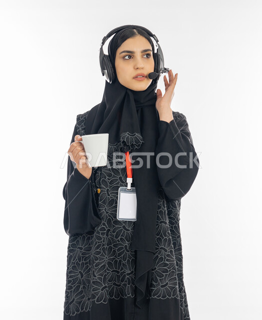 مهن ووظائف نسائية, النظر إلى شيء ما بتعمن وتركيز, بورتريه لإمرأة عربية خليجية اماراتية ترتدي العباءة وسماعة الاتصال تمسك بيدها كوب من القهوة, سيدة سعودية تعمل في مجال خدمة العملاء, الاجابة على أسئلة واستفسارات الزبائن, خلفية بيضاء