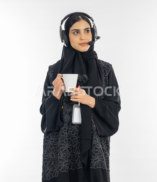 النظر إلى شيء ما بتعمن وتركيز, مهن ووظائف نسائية, بورتريه لإمرأة عربية خليجية اماراتية ترتدي العباءة وسماعة الاتصال تمسك بيدها كوب من القهوة, سيدة سعودية تعمل في مجال خدمة العملاء, الاجابة على أسئلة واستفسارات الزبائن, خلفية بيضاء