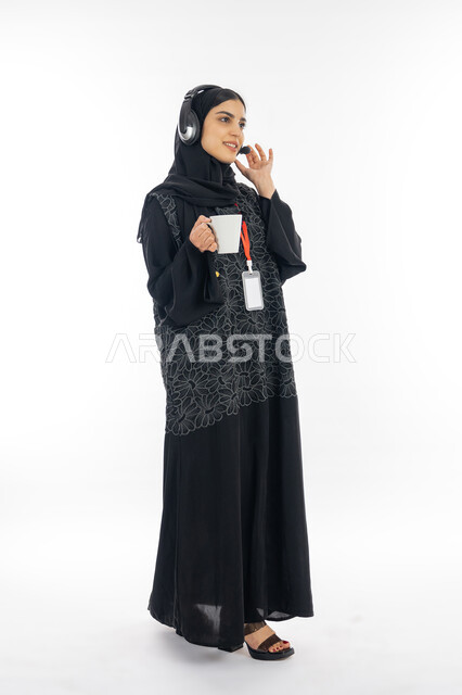 متابعة وتطوير العمل في الشركات الخليجية, بورتريه لإمرأة عربية خليجية إماراتية مبتسمة ترتدي العباءة وسماعة الاتصال تمسك كوب من القهوة، الرد على أسئلة واستفسارات الزبائن بإيماءات البشاشة, سيدة سعودية تعمل في مجال خدمة العملاء, صورة الجسم بالكامل, خلفية بيضاء